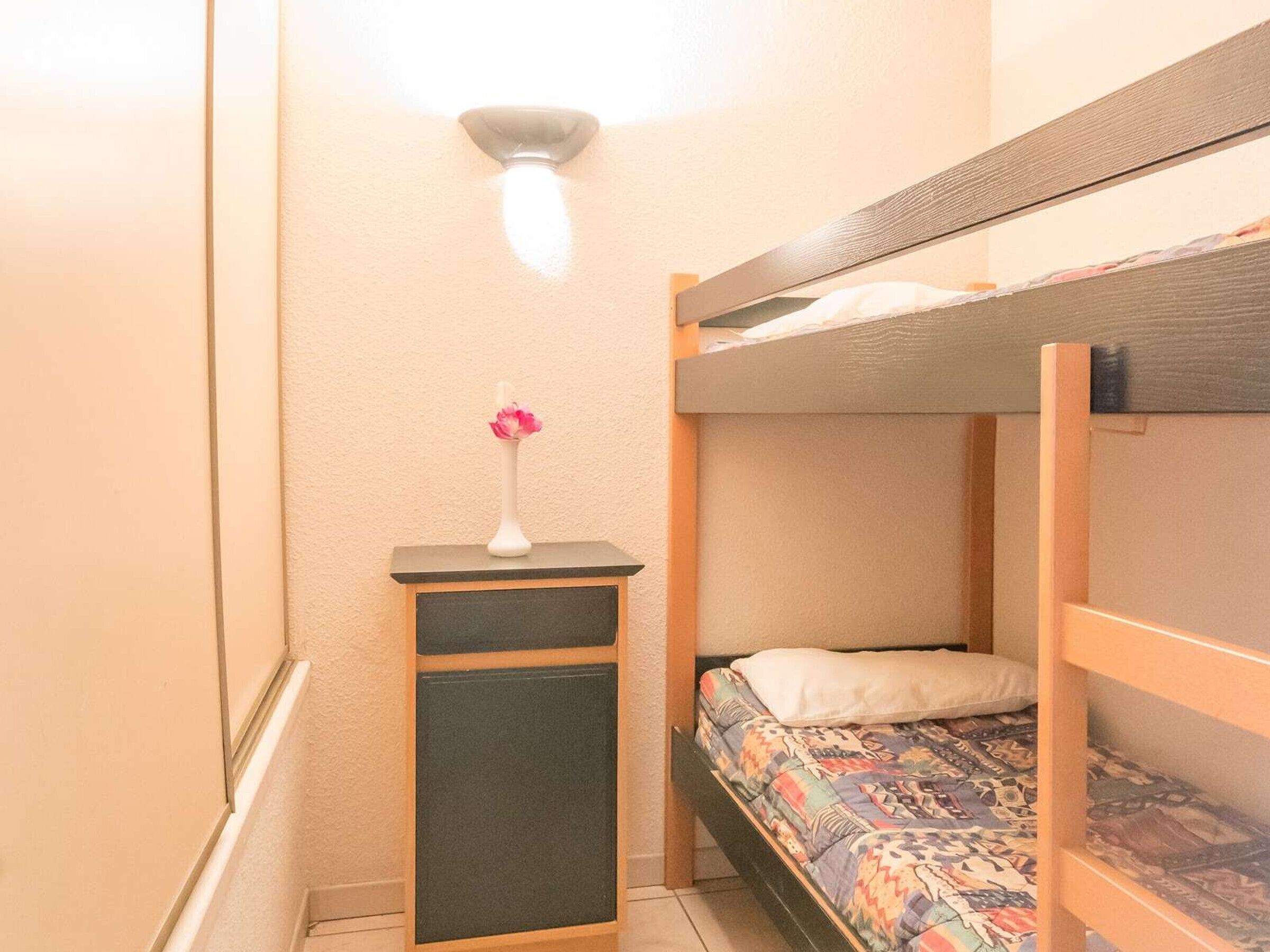 Et lille, kompakt værelse med køjeseng, et lille bord med en blomstervase og en vægmonteret lampe.
