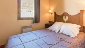 A bedroom with a bed, a nightstand, a wall-mounted lamp, and a window with a view of the outdoors.