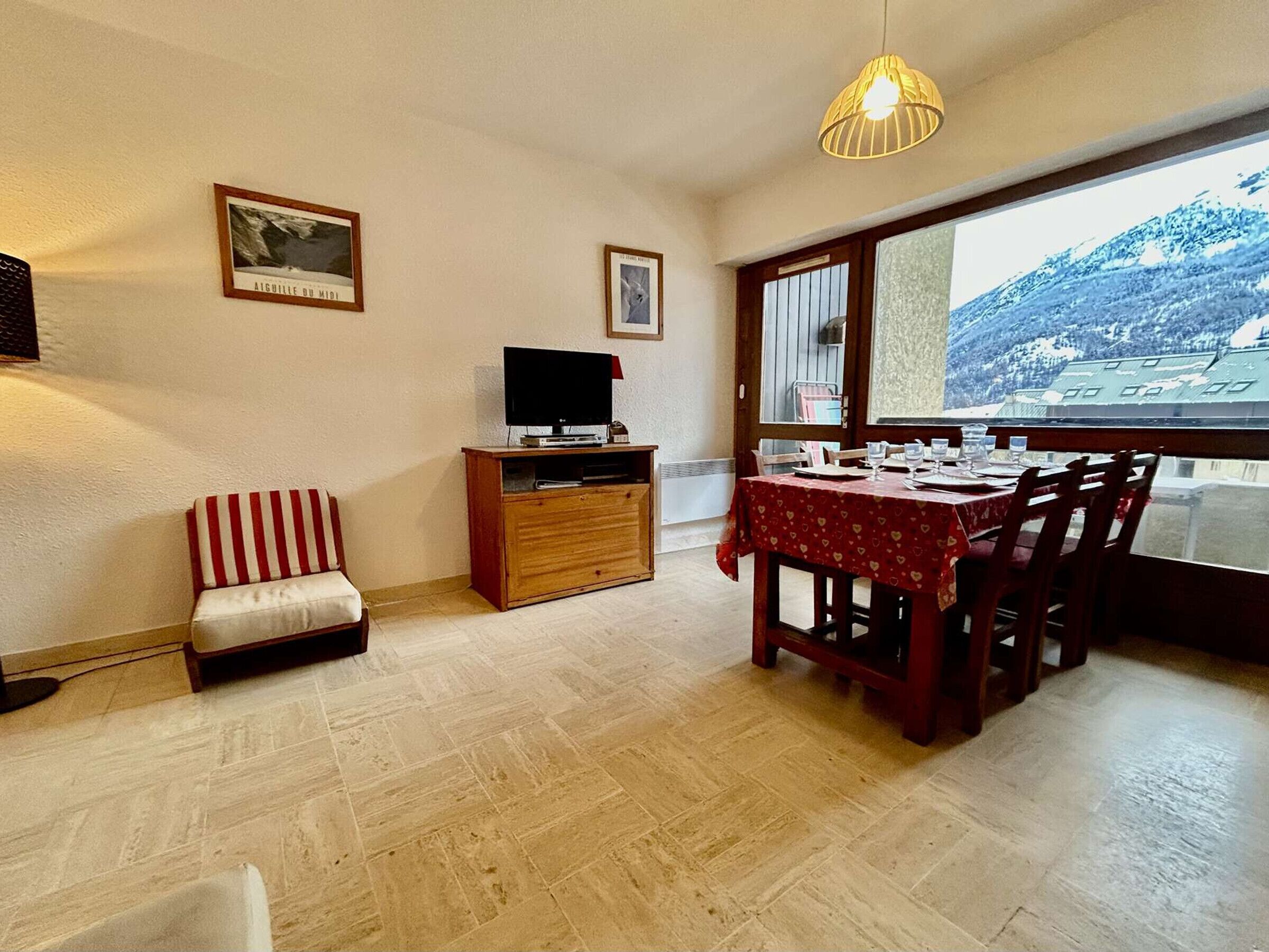 Un área de comedor con una mesa dispuesta para cuatro, un televisor sobre un mueble de madera, un banco acolchado y una ventana con vistas a la montaña.