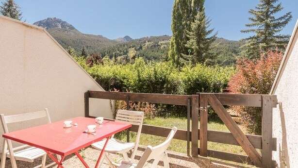 Un patio con una mesa roja y sillas blancas, con vistas a un viñedo y montañas.