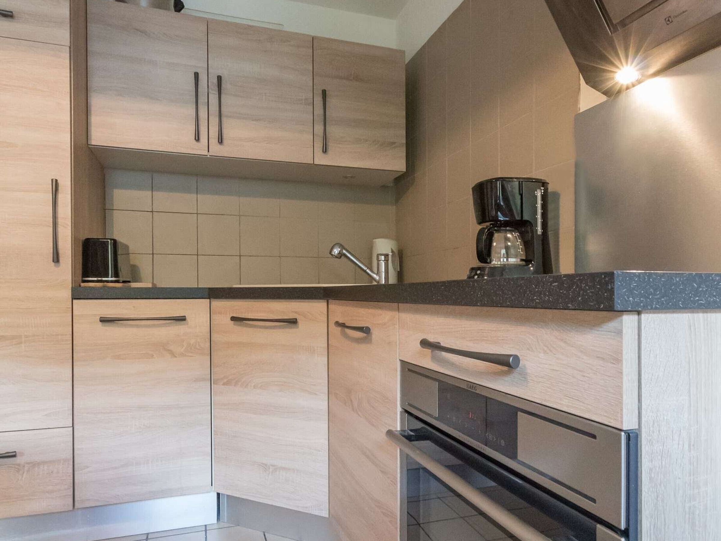 A modern kitchen with light wood cabinets, a black countertop, a coffee maker, and a stainless steel oven.