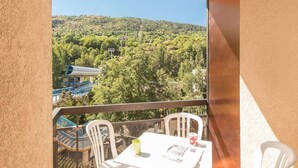A balcony with a table and chairs, overlooking a forested hillside and a building with a blue roof. - Night corner studio for 2/4 people in Briançon near slopes and shops (Briançon)