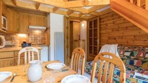 A wooden cabin interior with a dining table set for four, a kitchen area with wooden cabinets, and a staircase leading to an upper level.