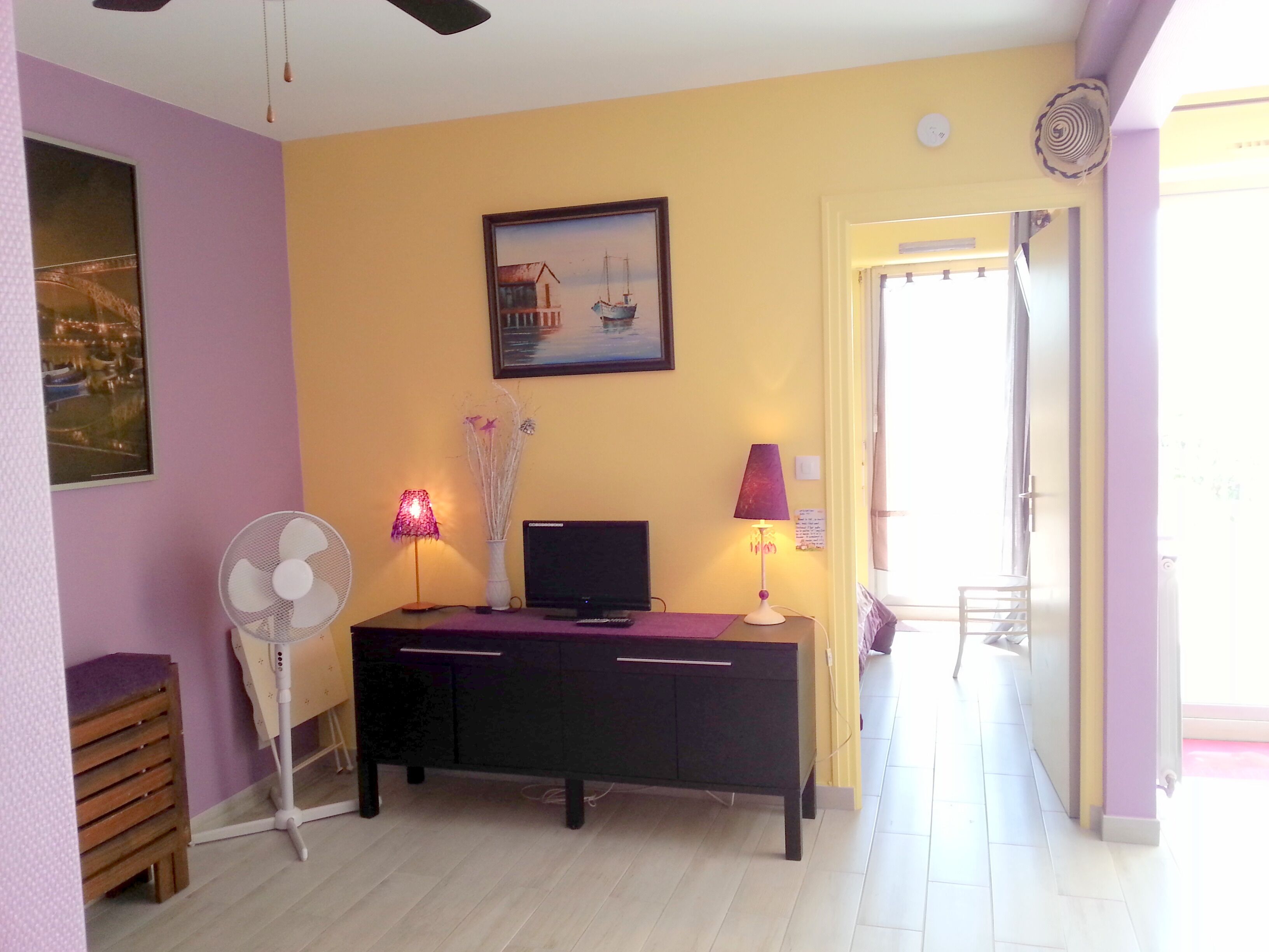 Une pièce comprenant un bureau, un ordinateur, un ventilateur et une lampe. Il y a un tableau au mur et une porte qui donne sur l’extérieur.