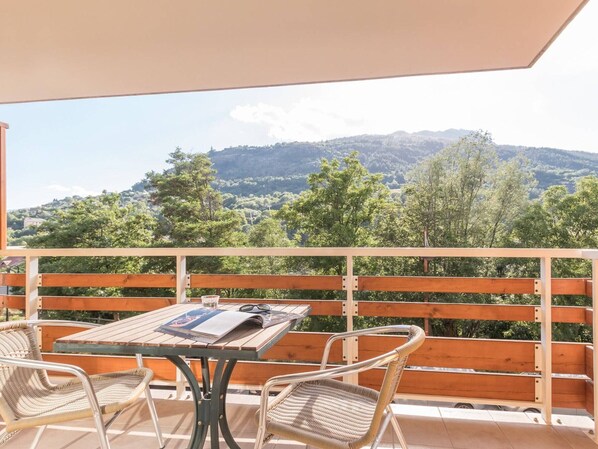 Un balcon avec une table en bois et deux chaises en osier, donnant sur un paysage montagneux.