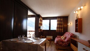 A cozy room with a wooden dining table set for two, a comfortable sofa, and a view of the mountains through large windows.