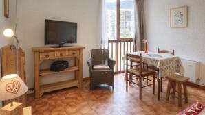 Ein gemütliches Zimmer mit Holzschreibtisch, Fernseher, Korbsessel, Esstisch mit Stühlen und einem Fenster mit Vorhängen.