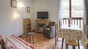 Ein gemütliches Zimmer mit Fernseher, Holzmöbeln, einem kleinen Tisch mit Blumenmuster-Tischdecke und einem Fenster mit Blick auf Gebäude.