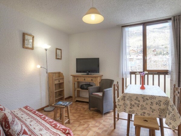 Une pièce confortable, dotée d  ’un téléviseur, de meubles en bois, d’une petite table recouverte d’une nappe à motifs floraux et d’une fenêtre donnant sur des immeubles.