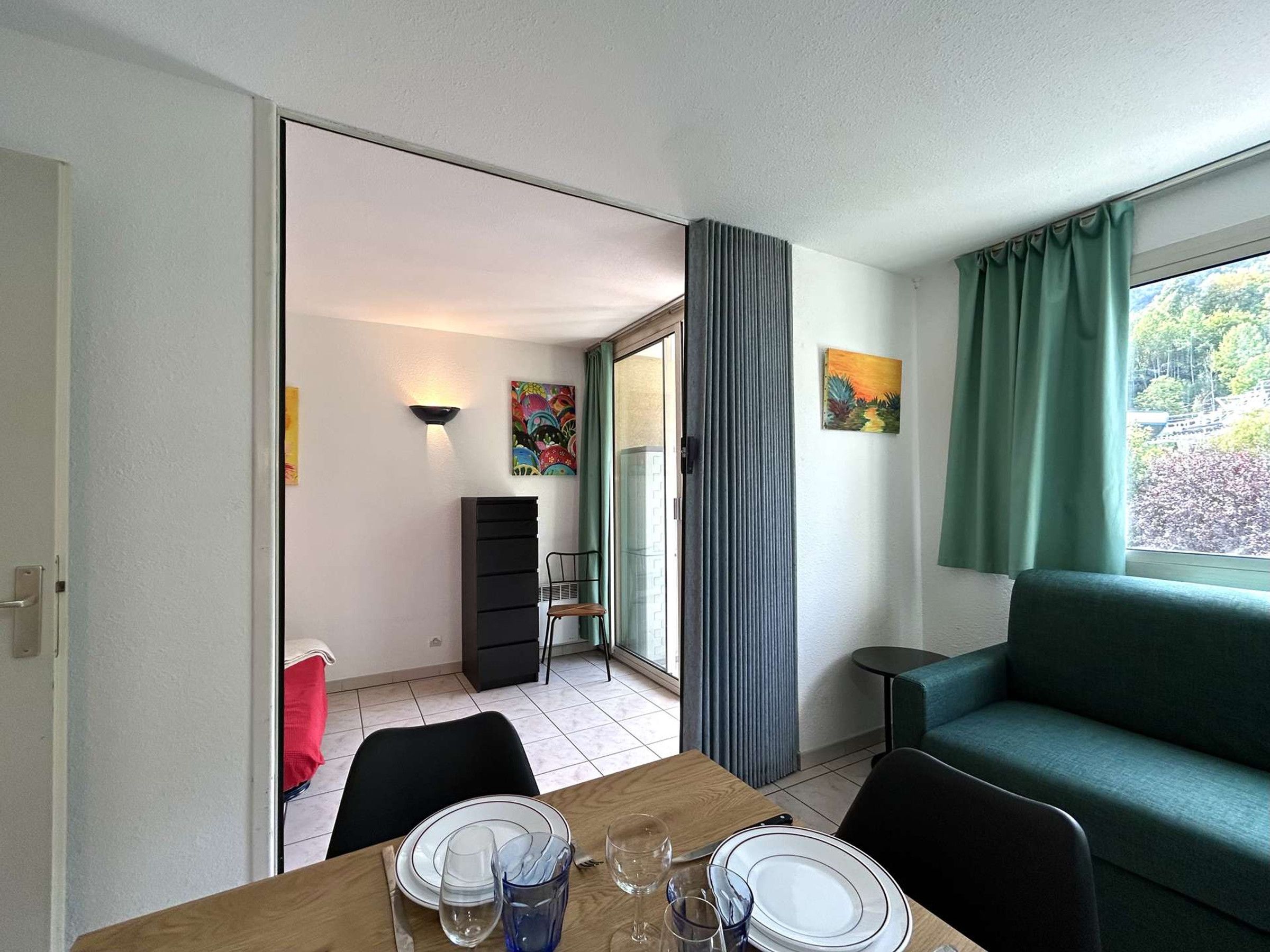 Un coin repas avec une table dressée pour deux personnes, un canapé vert et une vue par la fenêtre.