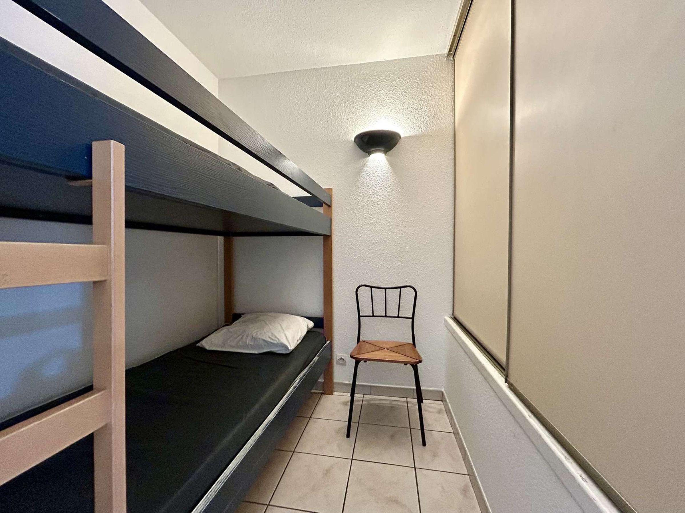 Une petite pièce étroite, avec un lit superposé, une seule chaise et un luminaire fixé au mur.