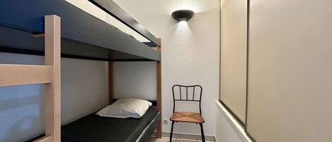 Une petite pièce étroite, avec un lit superposé, une seule chaise et un luminaire fixé au mur.