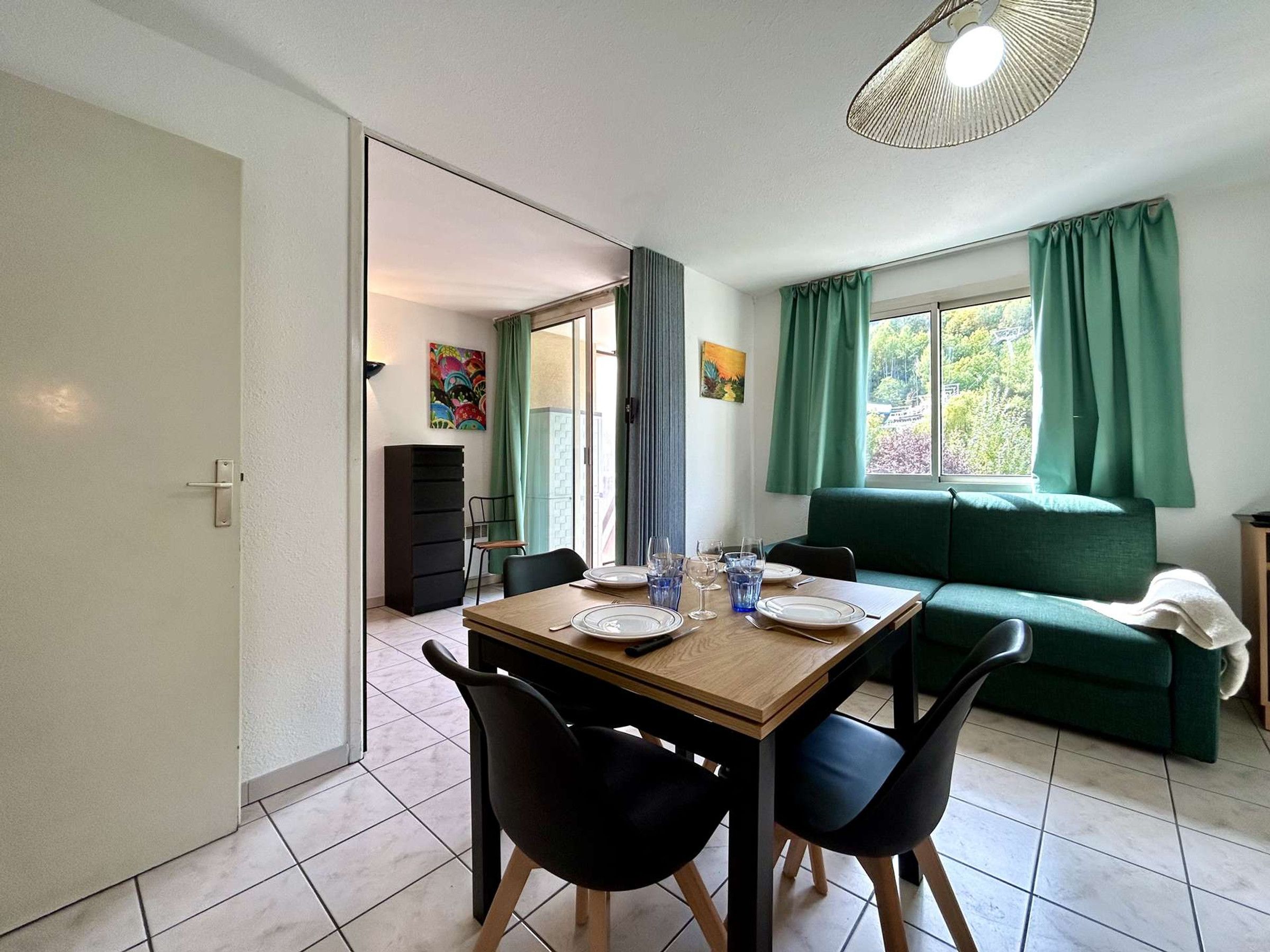 A dining area with a table set for two, a green sofa, and a view of the outdoors through a window.