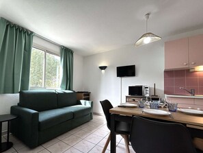 Una sala moderna con un sofá verde, una mesa de comedor preparada para dos personas, un televisor de pantalla plana montado en la pared y una zona de cocina con armarios rosados.