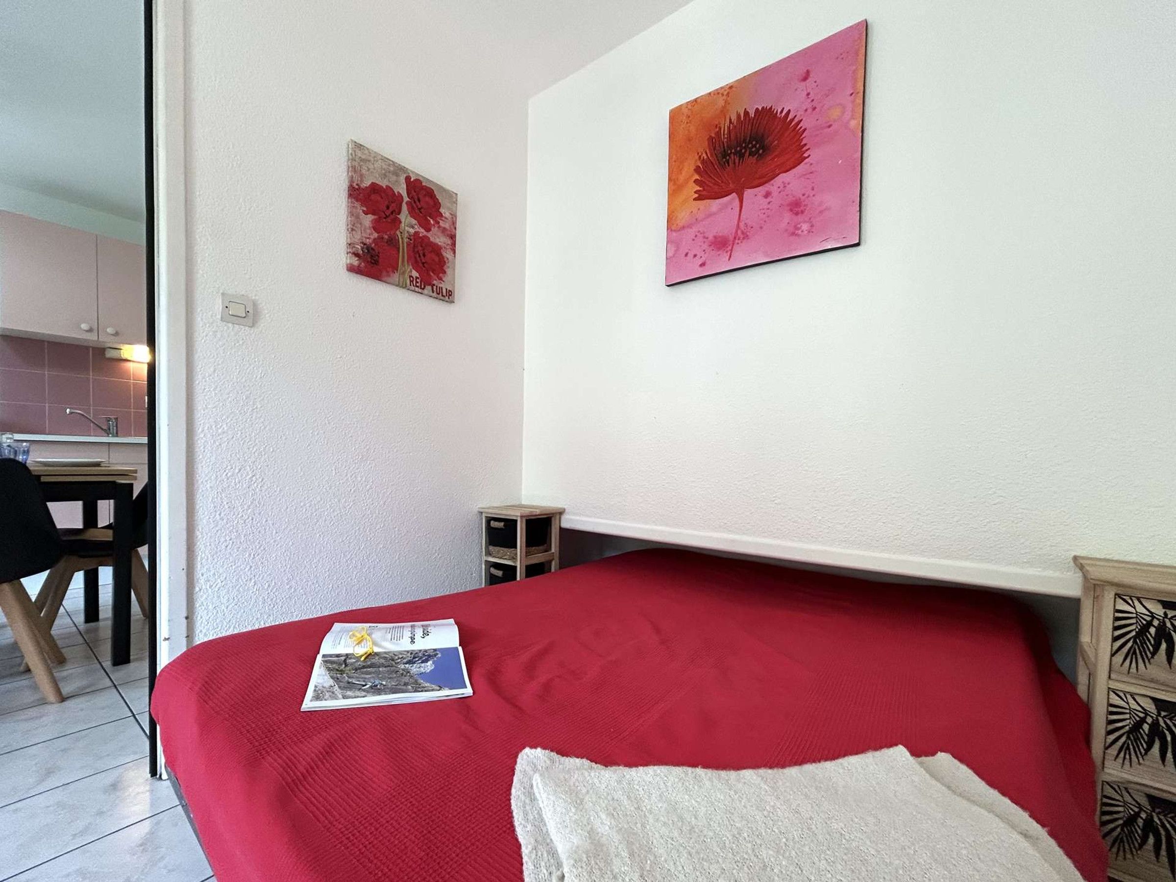 A room with a bed, a small table, a chair, and two paintings on the wall.