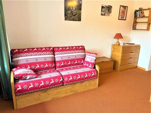 En soffa i trä med ett rött och vitt mönstrat överdrag, ett litet bord i trä med en lampa och en bokhylla placerad mot en enkel vägg.