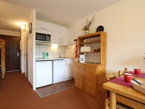 Ett kompakt kök med vita skåp, en mikrovågsugn, en kaffebryggare och ett träskåp med hyllor.
