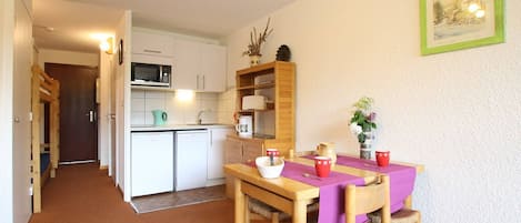 Une cuisine compacte avec des meubles blancs, un four à micro-ondes et un évier. On y trouve une table à manger recouverte d’une nappe violette et agrémentée de tasses rouges.