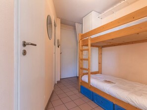 Um corredor estreito com uma cama de solteiro de madeira e uma porta com um espelho redondo e um interruptor de luz.