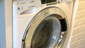 Una lavadora de carga frontal con un panel de control, ubicada en una habitación con piso de baldosas y pared de ladrillo blanco.