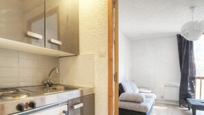 Une cuisine compacte dotée d’un four encastré et d’un évier, attenante à un espace de vie comprenant un canapé et une fenêtre.