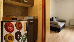 Une cuisine compacte, agrémentée d’un réfrigérateur décoré de capsules de bouteilles, d’un four encastré et d’un évier. À côté se trouve un espace salon avec un canapé et une petite table.