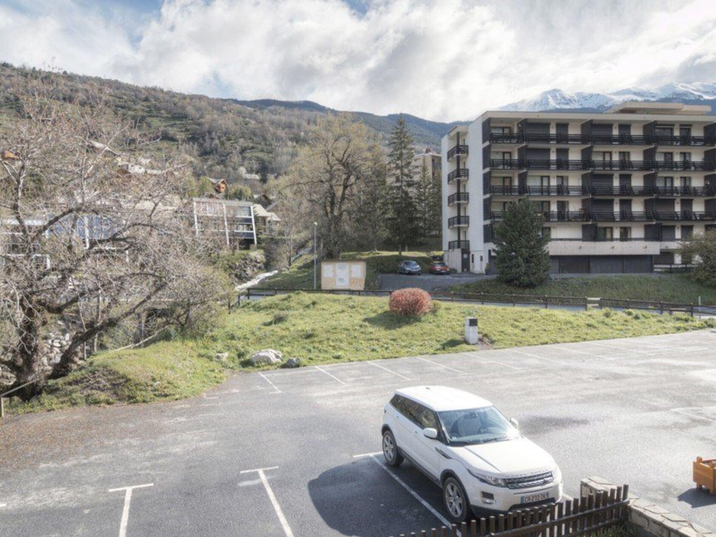 Un espace de stationnement avec un VUS blanc, un immeuble à plusieurs étages doté de balcons et un paysage montagneux en arrière-plan.