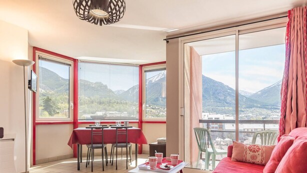 A room with a dining table and chairs, a sofa, and large windows offering a mountain view. - 2-room apartment with balcony, near slopes - Briançon (Briançon)