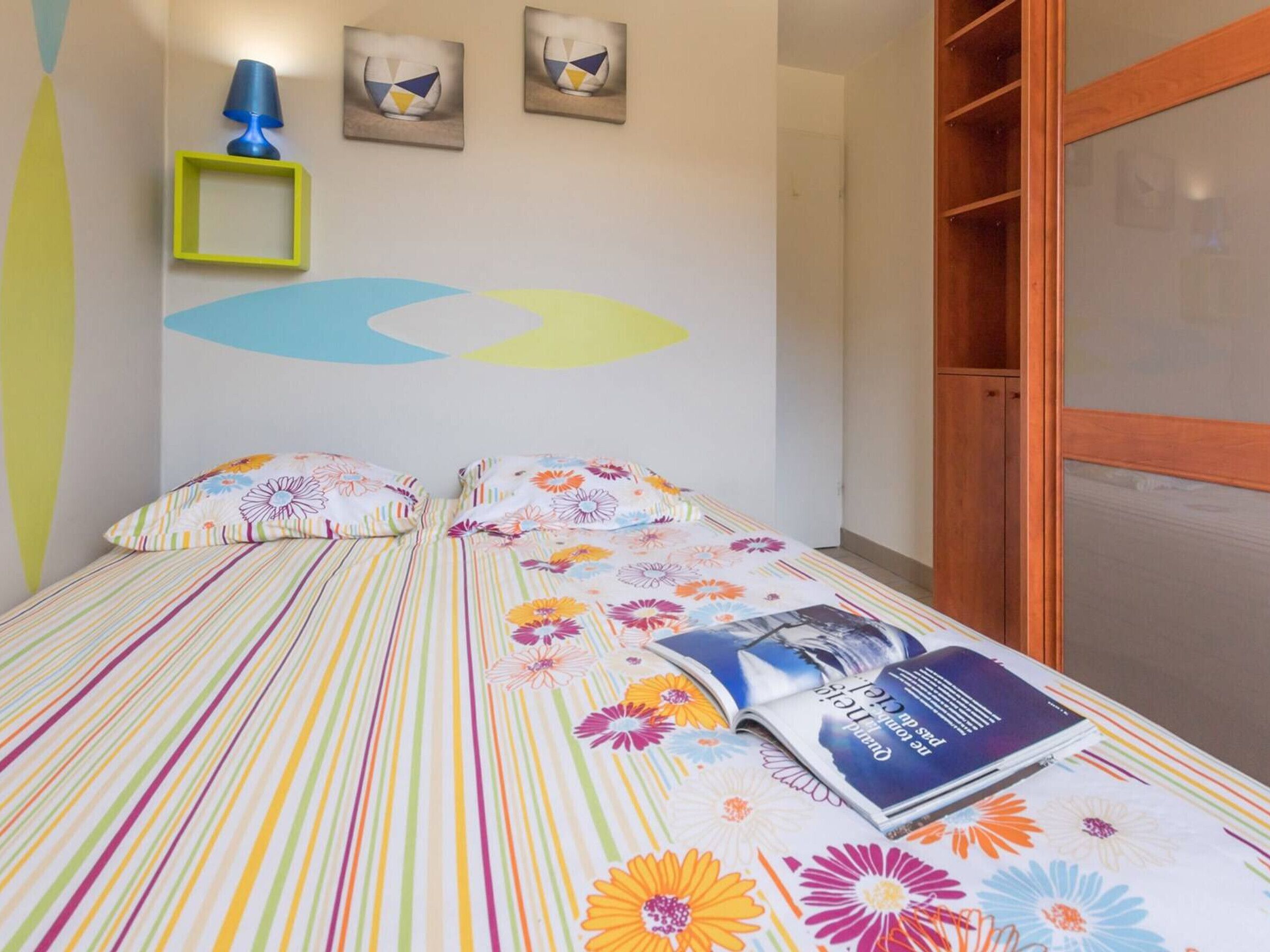 Een bed met een gestreept dekbedovertrek en bloemenkussens, een blauwe lamp en twee ingelijste foto's aan de muur.