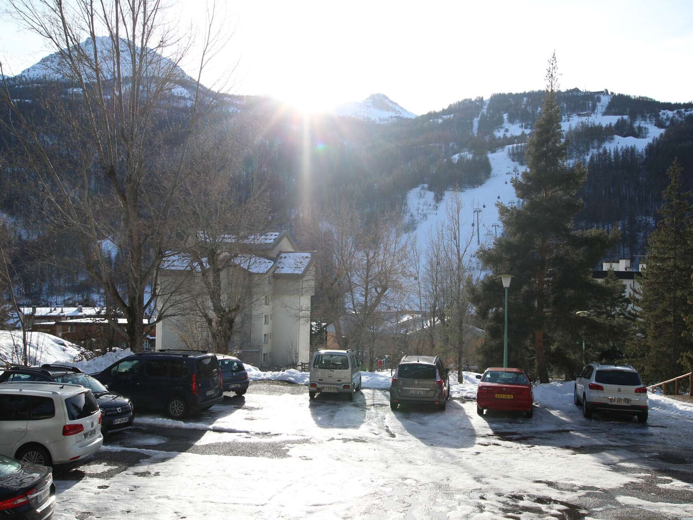 A parking lot with several cars, a building with a sloped roof, and snow-covered mountains in the background.