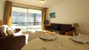A cozy living room with a sofa, a dining table set for two, and a large window offering a view of a snowy landscape. - Cozy 6-Person Apartment with Mountain View and Balcony in Serre-Chevalier (Saint-Chaffrey)