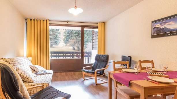 A cozy room with a sofa, two chairs, a dining table with a pink tablecloth, and a large window with curtains.