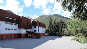 A building with a white facade and brown roof, labeled "L'IZOARD," with a parking area in front and a mountainous backdrop.