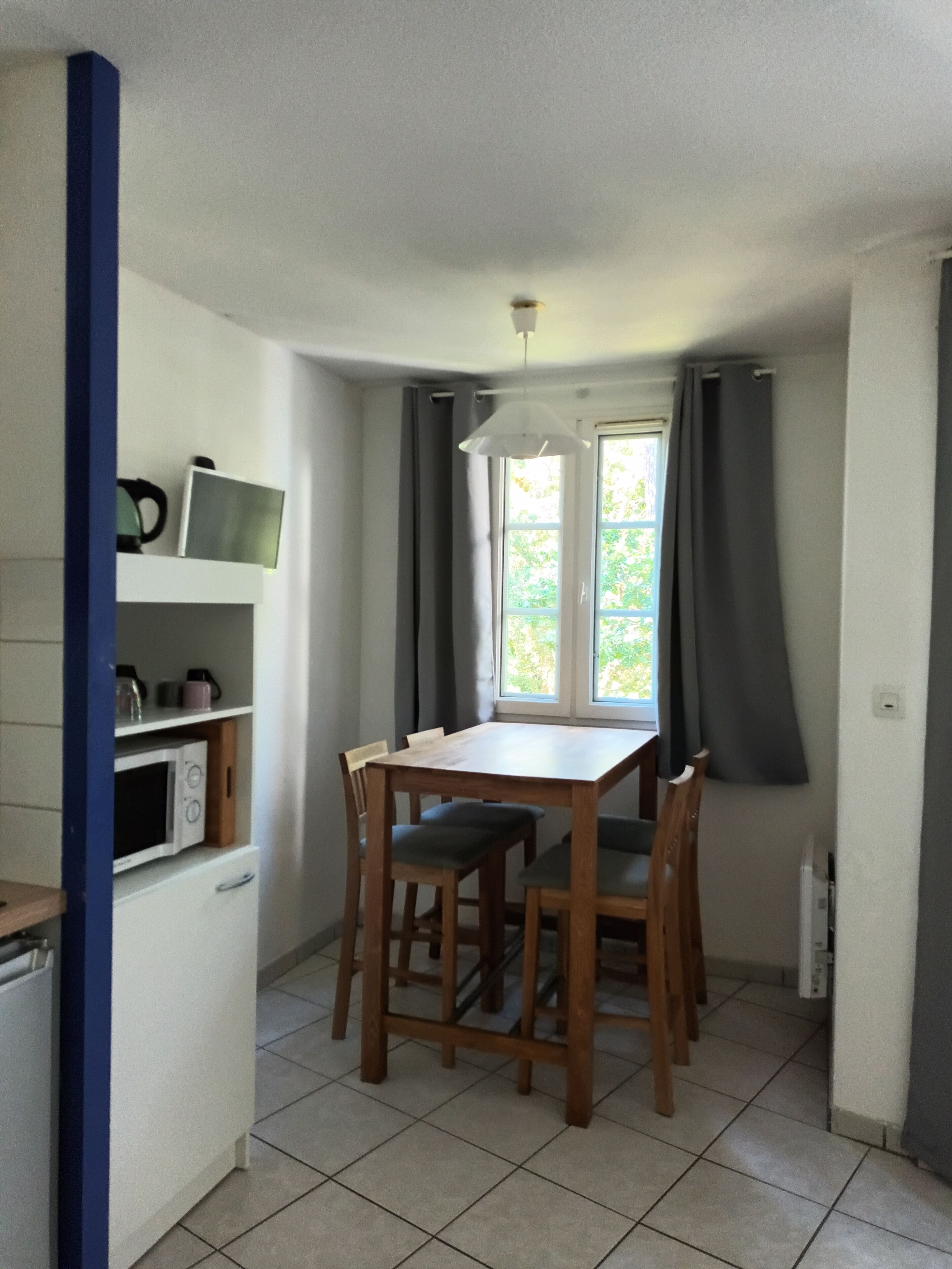A small, bright kitchen with a dining table and chairs, a microwave, and a window with curtains.