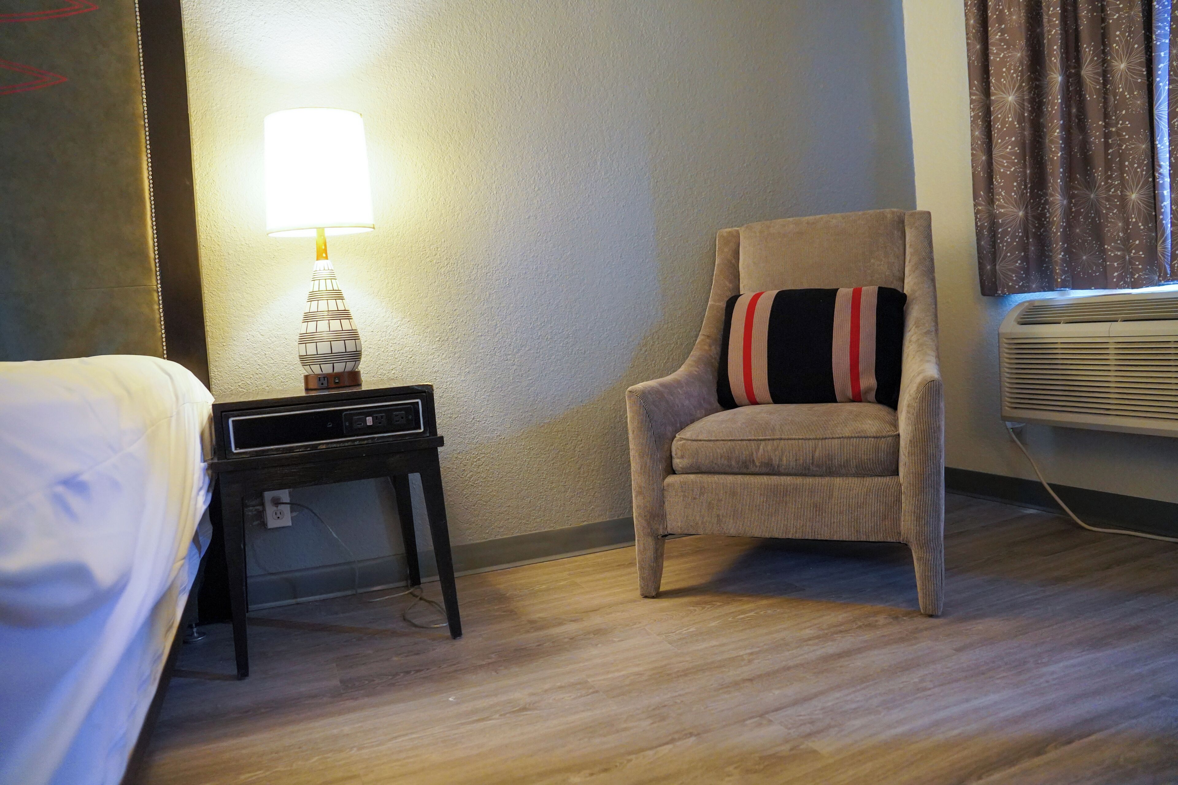 A hotel room with a bed, a chair with a striped pillow, a bedside table with a lamp, and an air conditioner.