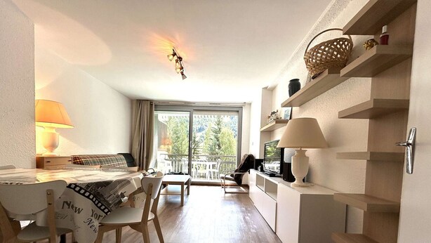 A modern, well-lit room with a dining area, a sofa, and a balcony with a view of greenery.