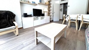 A modern living room with a flat-screen TV, white cabinets, a wooden coffee table, and a dining area with a white table and chairs.