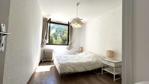 A bedroom with a bed, a bedside table, a lamp, a window with curtains, and a radiator.