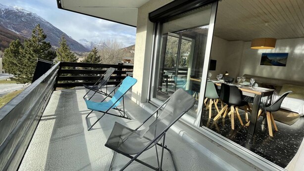 A balcony with two chairs, a dining area with a table and chairs, and a view of mountains.