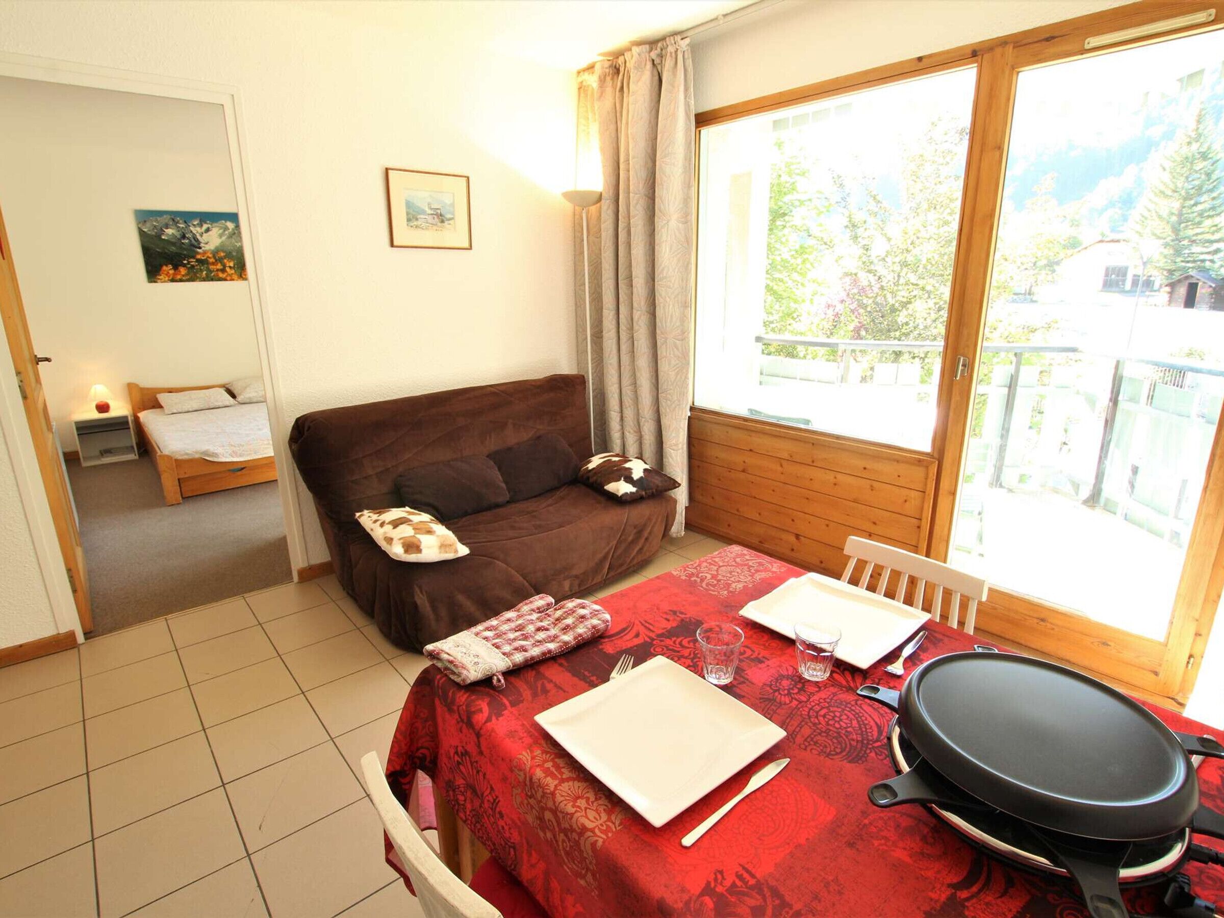 A cozy room with a bed, a sofa, a dining table set for two, and a large window with a view of greenery.