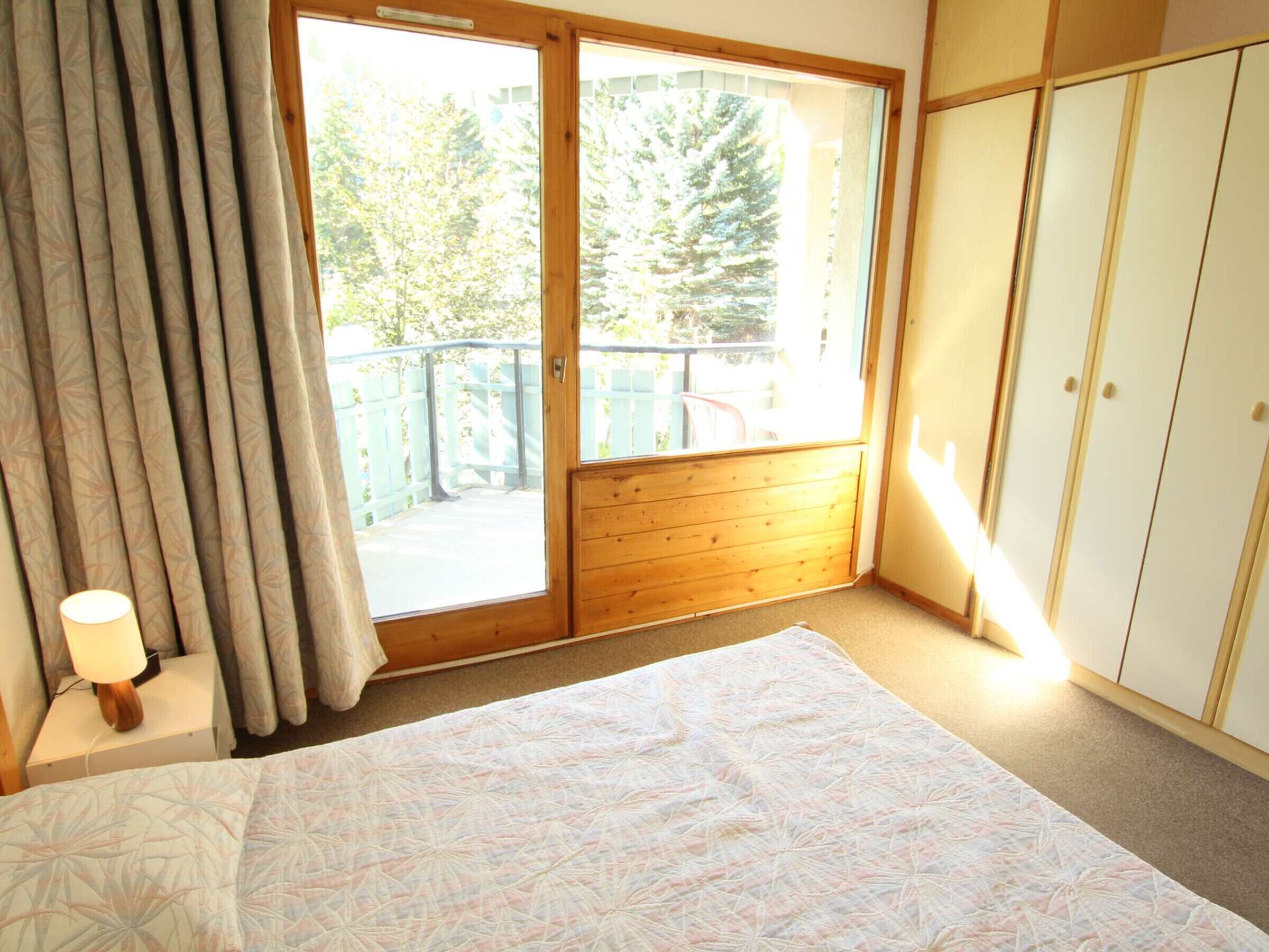 A bedroom with a bed, a nightstand, a lamp, a window with curtains, and a wardrobe.