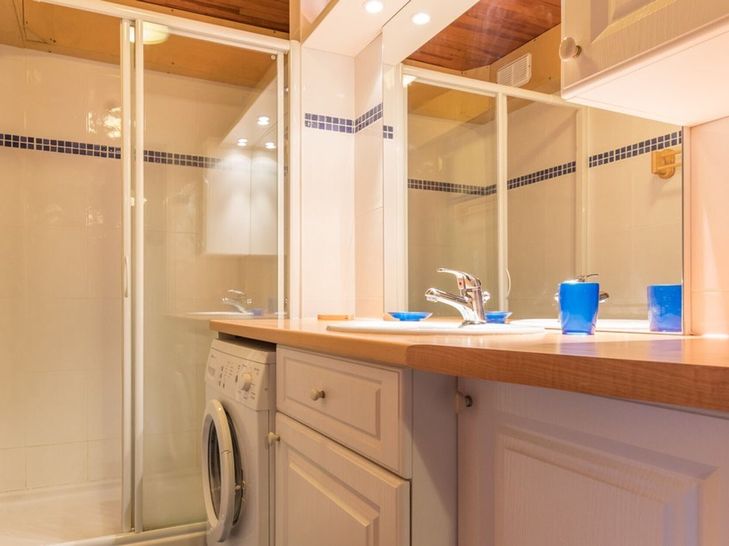 A modern bathroom with a glass shower enclosure, a built-in washing machine, and a countertop with a sink and blue cups.