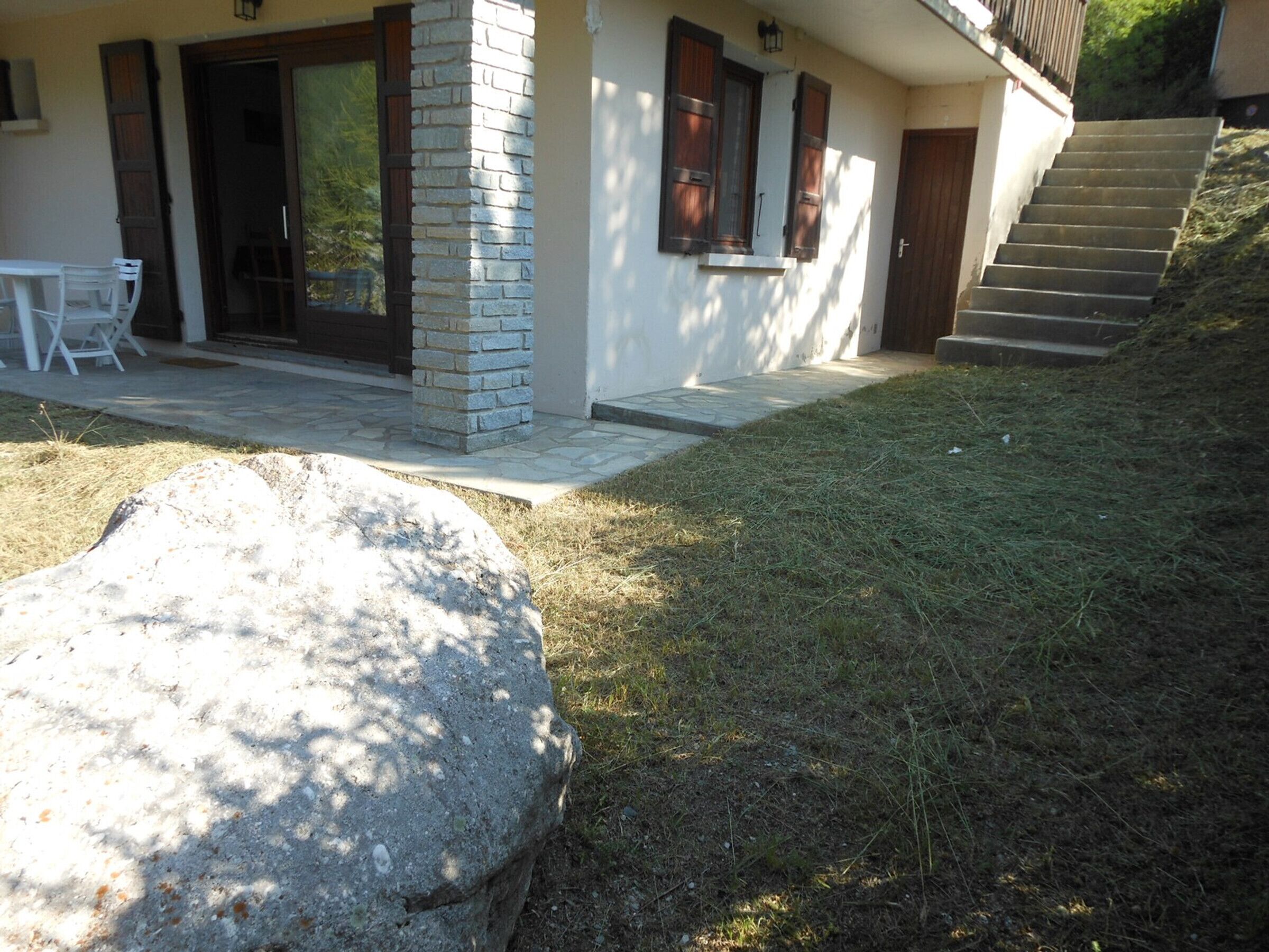 Une maison moderne avec un pilier en pierre, un patio avec une table et des chaises, et un escalier menant à l’entrée.