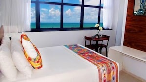 Ein Hotelzimmer mit großem Fenster und Meerblick, einem Bett mit weißer und farbenfroher Bettwäsche, einem kleinen Tisch mit Blumenvase und einer Wand mit Holzelementen.