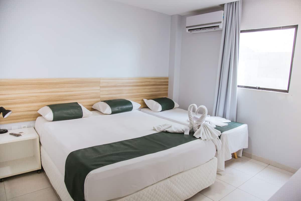 A hotel room with a large bed, a bedside table, an air conditioning unit, and a window with curtains.
