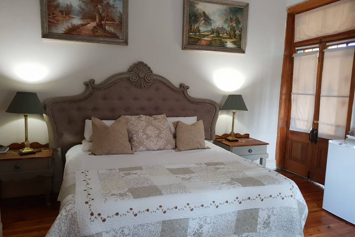 Un dormitorio ordenado con una cama grande, dos mesitas de noche con lámparas, una puerta de madera y dos cuadros de paisajes en la pared.