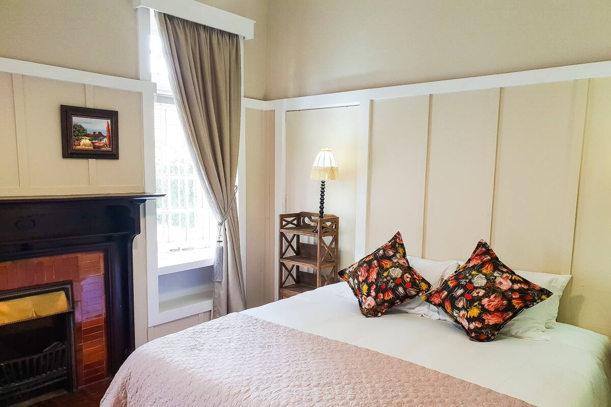 Un dormitorio con cama, chimenea, ventana con cortinas y lámpara.