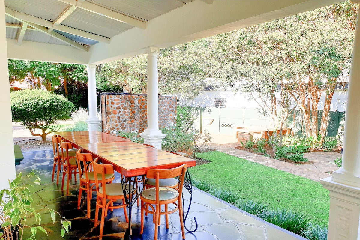 Un porche cubierto con una mesa de madera y sillas color naranja, rodeado por un muro de piedra y un césped cuidado.