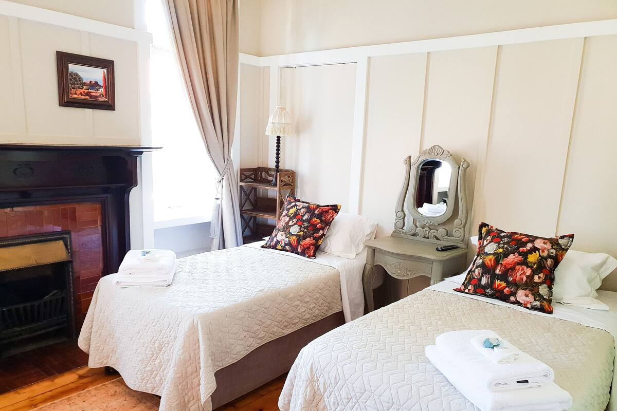 Habitación de hotel con dos camas, una chimenea, un espejo y un ventanal con cortinas.