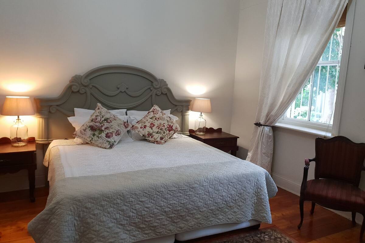 Una cama bien hecha con almohadones florales, dos lámparas de noche y una ventana con cortinas.